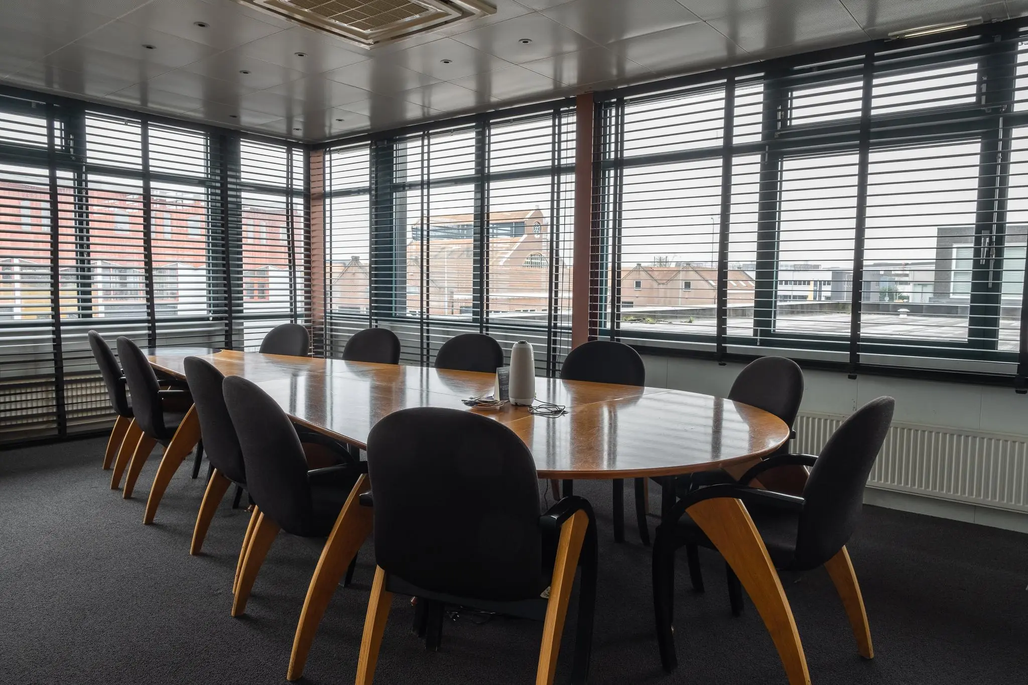 Vergaderruimte met ovale houten tafel en zwarte stoelen aan de Johan van Hasseltweg, omgeven door ramen met jaloezieën.